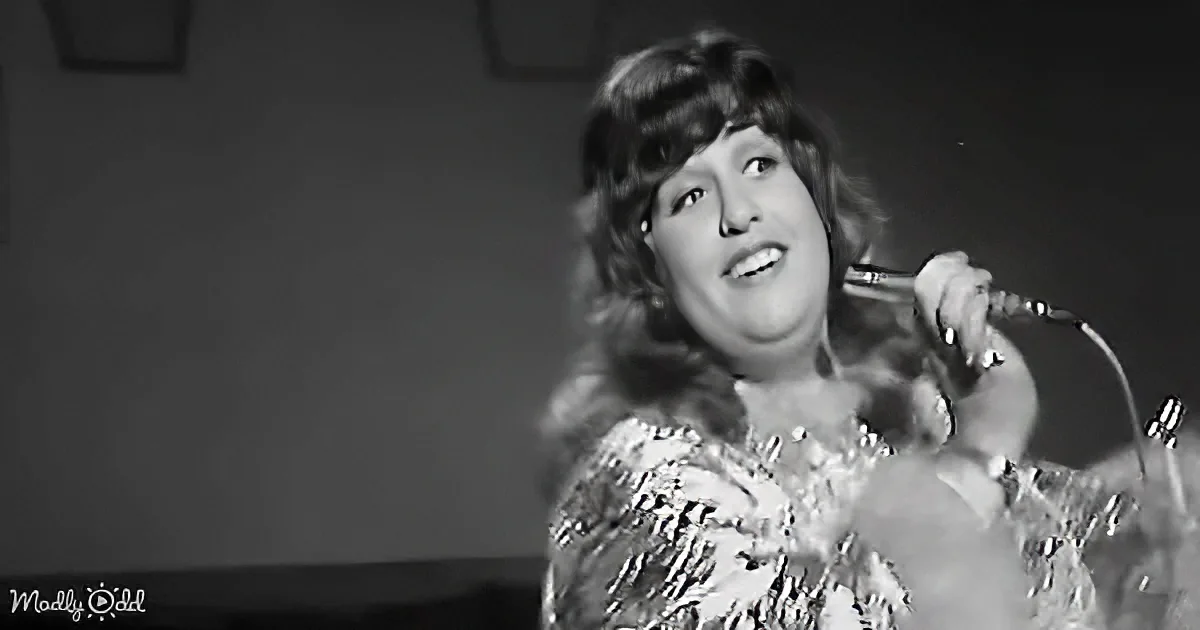 A woman with curly hair, wearing a sparkly dress, sings into a microphone.