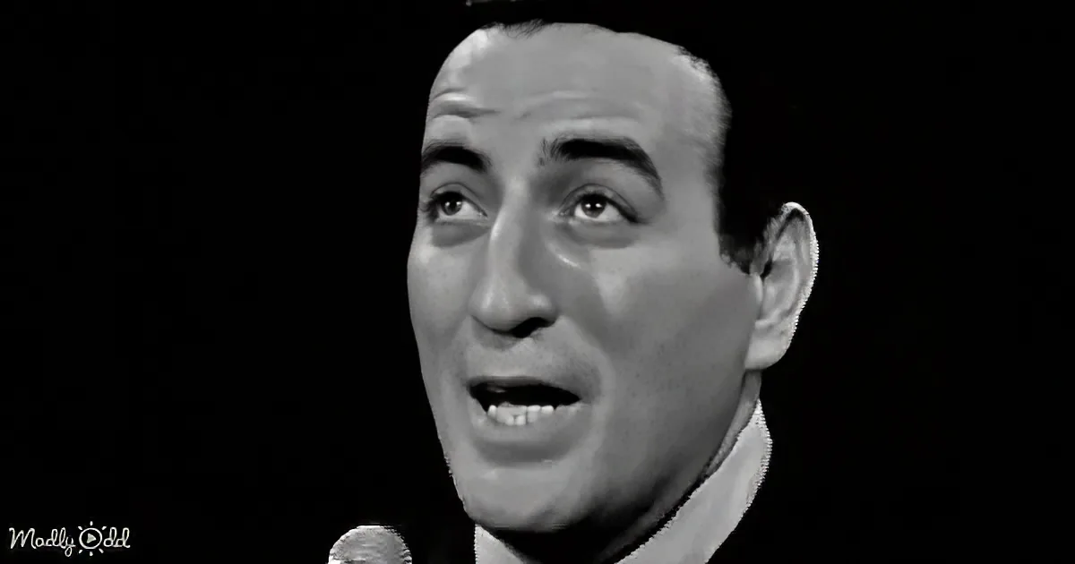 A man singing into a microphone, wearing a white shirt, against a dark background.