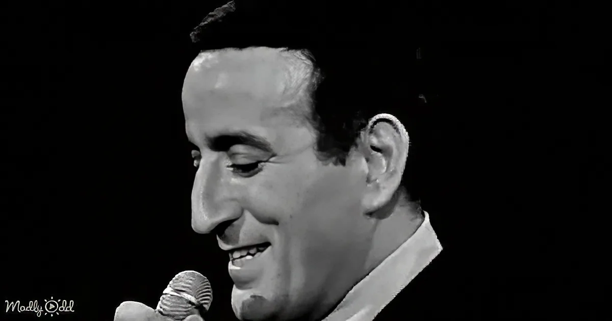 A man smiles while holding a microphone, wearing a white shirt, against a dark background.