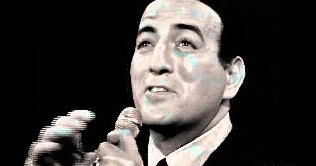 A smiling man holding a microphone, wearing a suit, against a dark background.