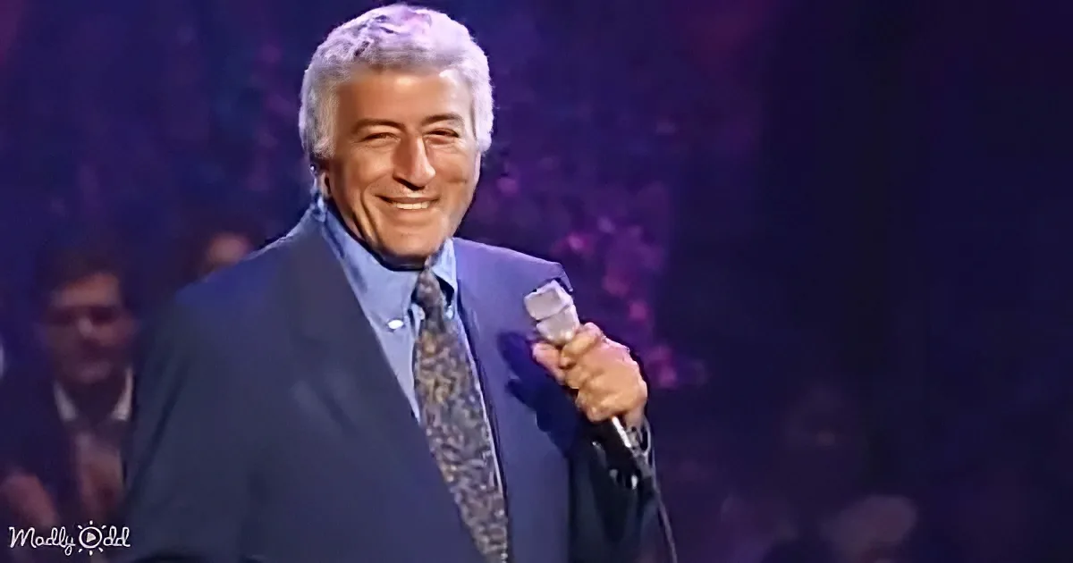 A man in a blue suit and patterned tie holds a microphone, smiling on stage with a dark background.