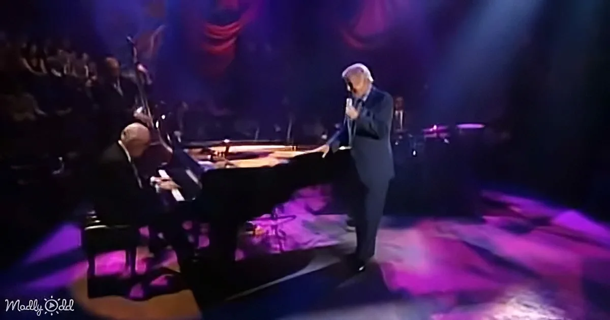 A man in a suit sings into a microphone on stage, accompanied by a pianist.