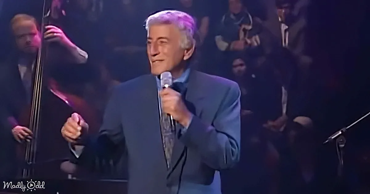 A man in a suit sings into a microphone on stage, surrounded by musicians.