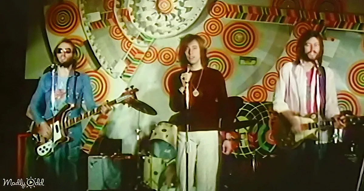Three musicians perform on stage with psychedelic background, wearing colorful outfits.