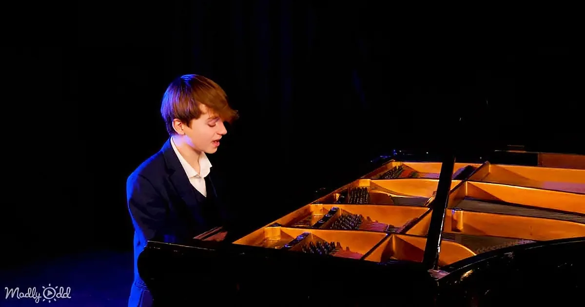 A young boy in a suit plays a grand piano under stage lighting.