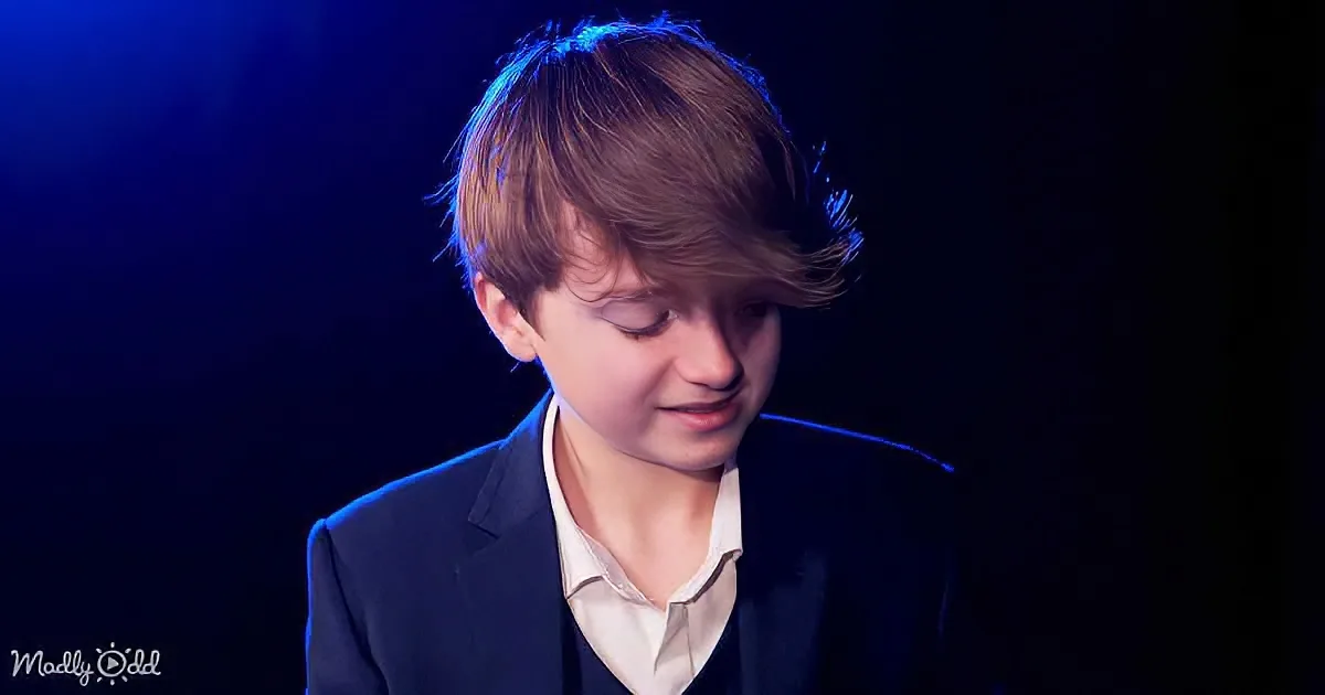 A young boy in a suit and white shirt, eyes closed, head slightly tilted, against a dark background with blue lighting.