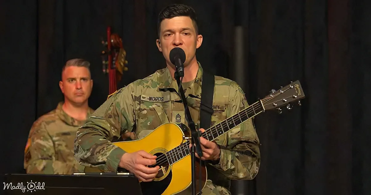 A soldier in camouflage plays guitar and sings into a microphone, accompanied by another soldier in the background.