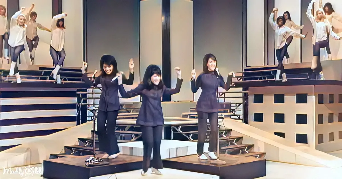 Three women in black outfits perform on a stage with a backdrop of a cityscape and a rooftop scene.