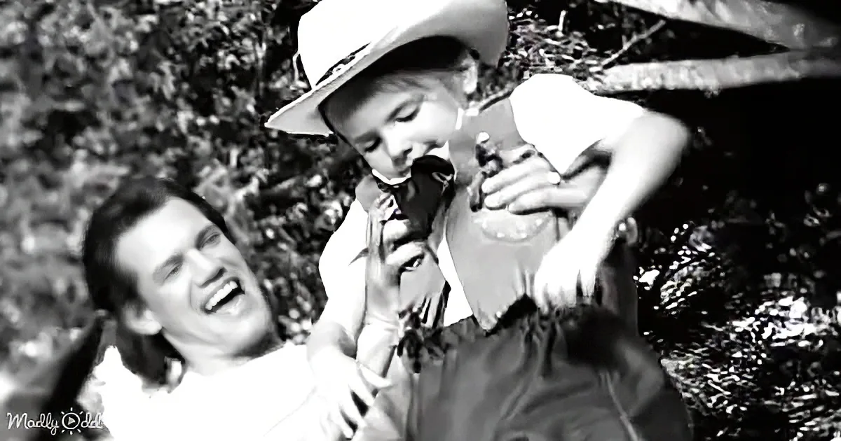A woman in a cowboy hat holds a bottle, smiling, with a man in the background.