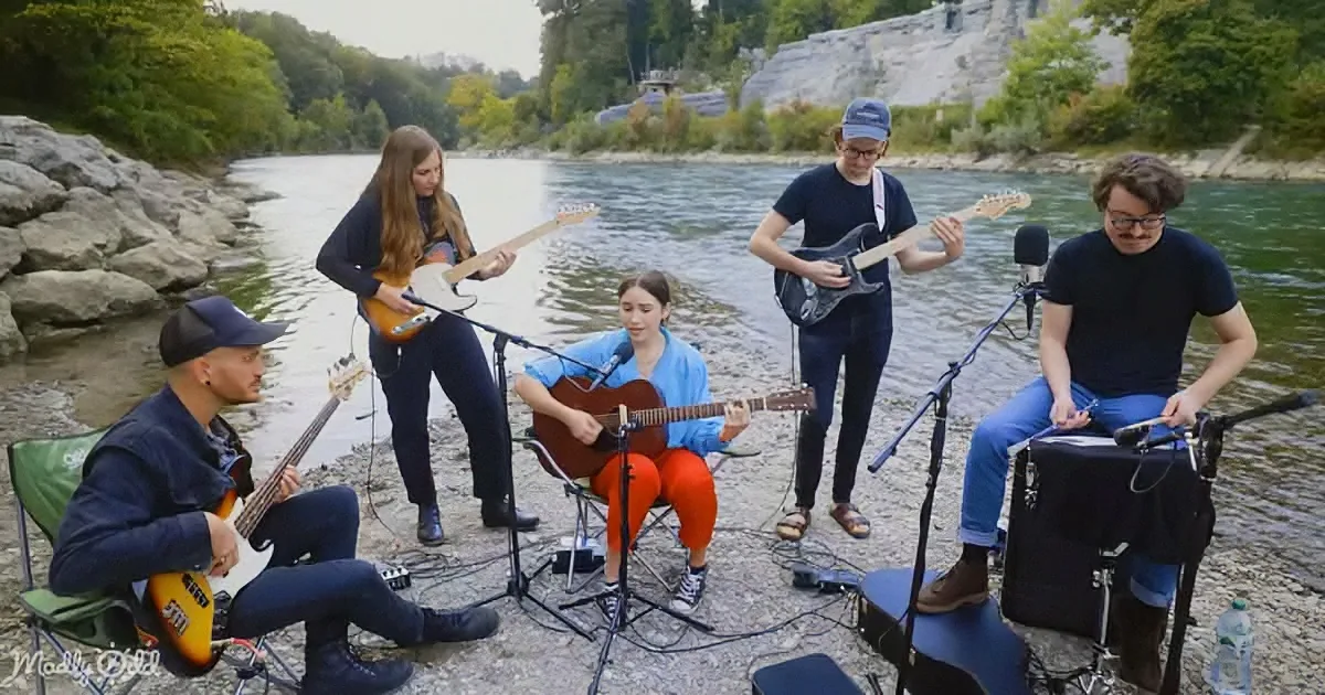 A group of musicians plays music outdoors by a river, with microphones and instruments.