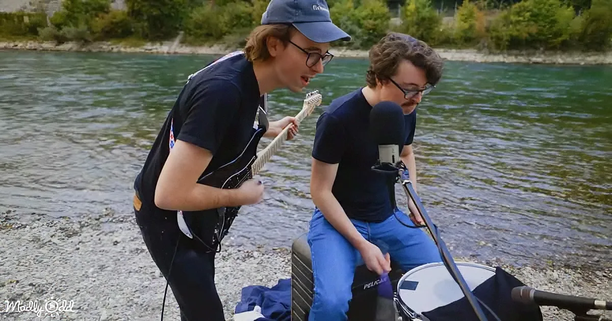 Two individuals play music outdoors near a body of water, one strumming guitar, the other singing into microphone.