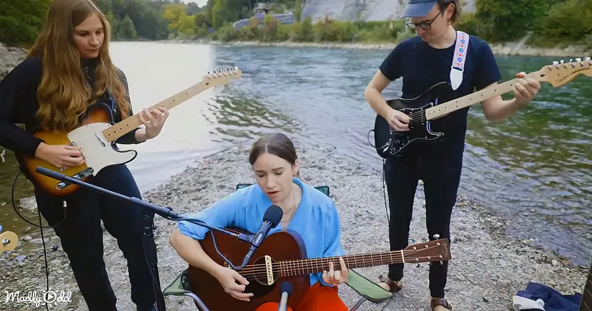 A woman in blue plays an acoustic guitar near a river, accompanied by two musicians with electric guitars.