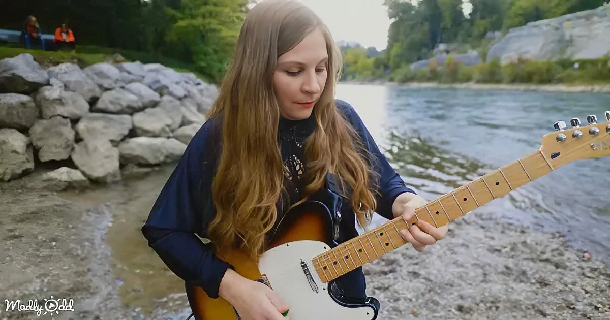 A woman with long hair plays a guitar by a river, surrounded by rocks and trees.