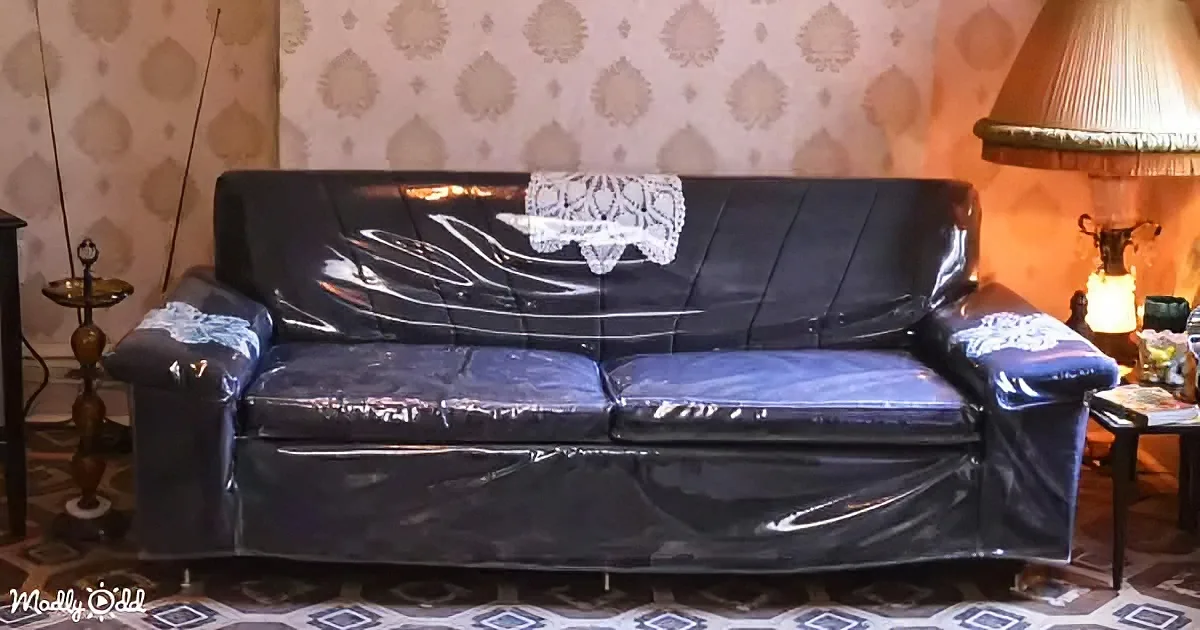 A black sofa covered in plastic with a lace doily on top, placed in a room with patterned wallpaper and a lamp.