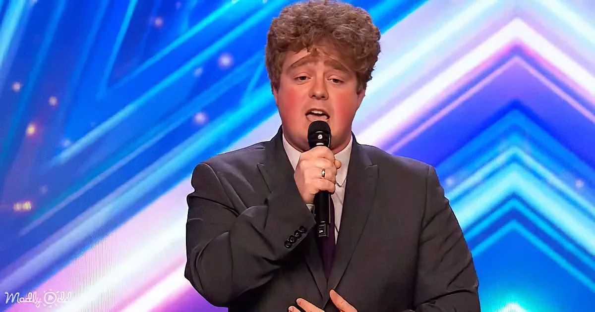 A man in a suit sings into a microphone on a stage with a blue, geometric background.
