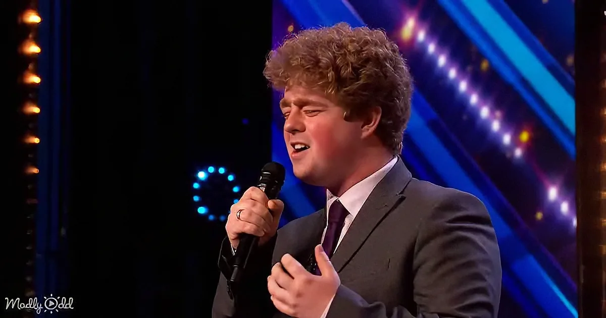 A man in a suit sings into a microphone on a stage with blue lights.