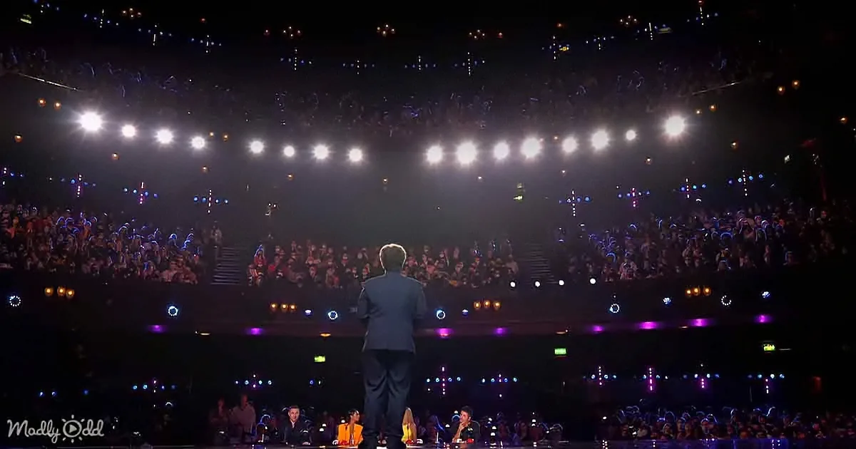 A man in a suit stands on a stage facing a large, illuminated audience in a dark arena.
