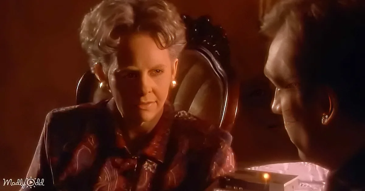 A person with curly hair, wearing a red shirt, sits at a table with another person, both in a dimly lit setting.