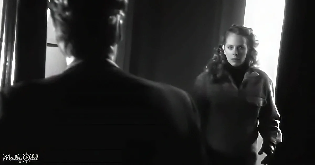 A woman in a dark coat stands in a doorway, facing a man in a suit, in a dimly lit room.