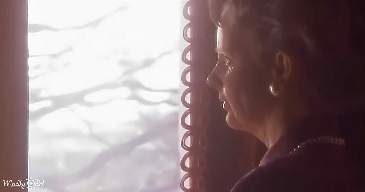 A woman with short hair, wearing a necklace, looks out a window with a curtain, bathed in sunlight.