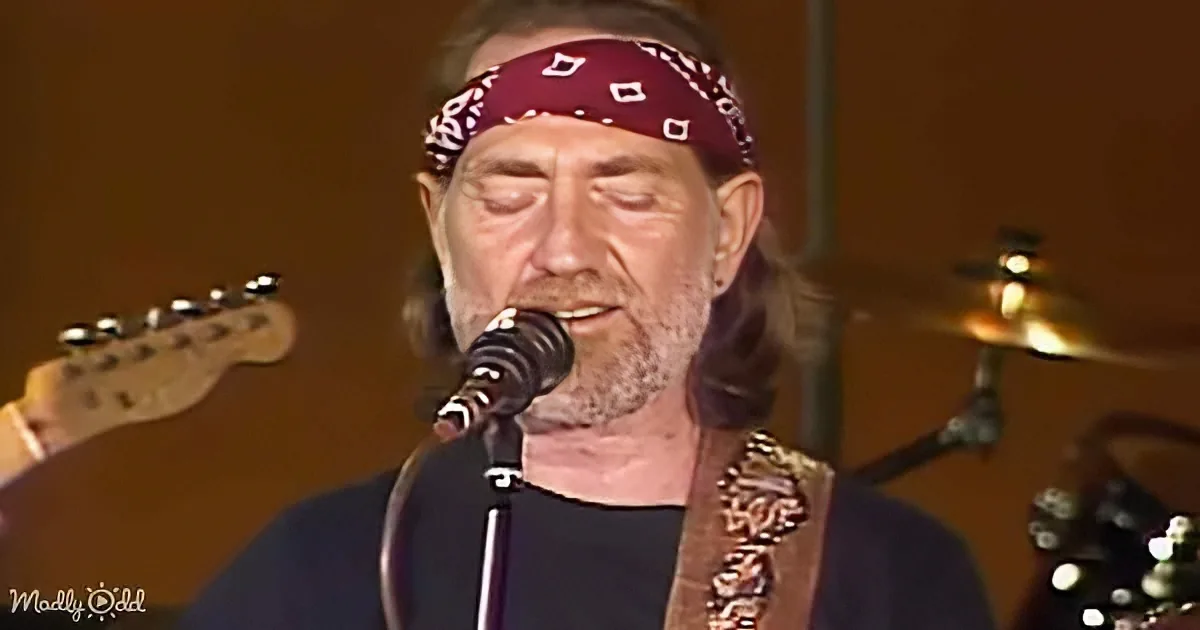 A man with a bandana and beard sings into a microphone, holding a guitar, with a drum set in the background.