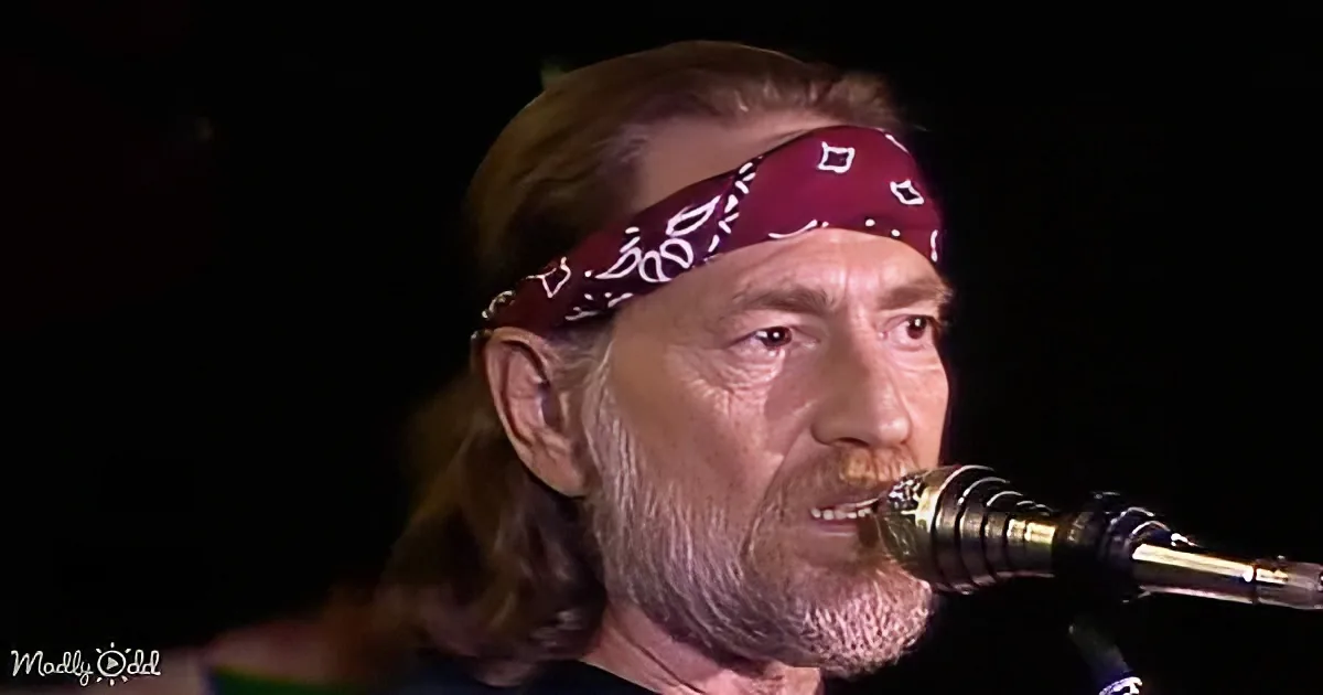 A man with a bandana and long hair sings into a microphone against a dark background.