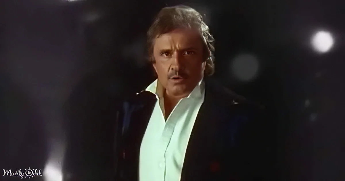 A man in a white shirt and black jacket stands against a dark background with a microphone in front of him.