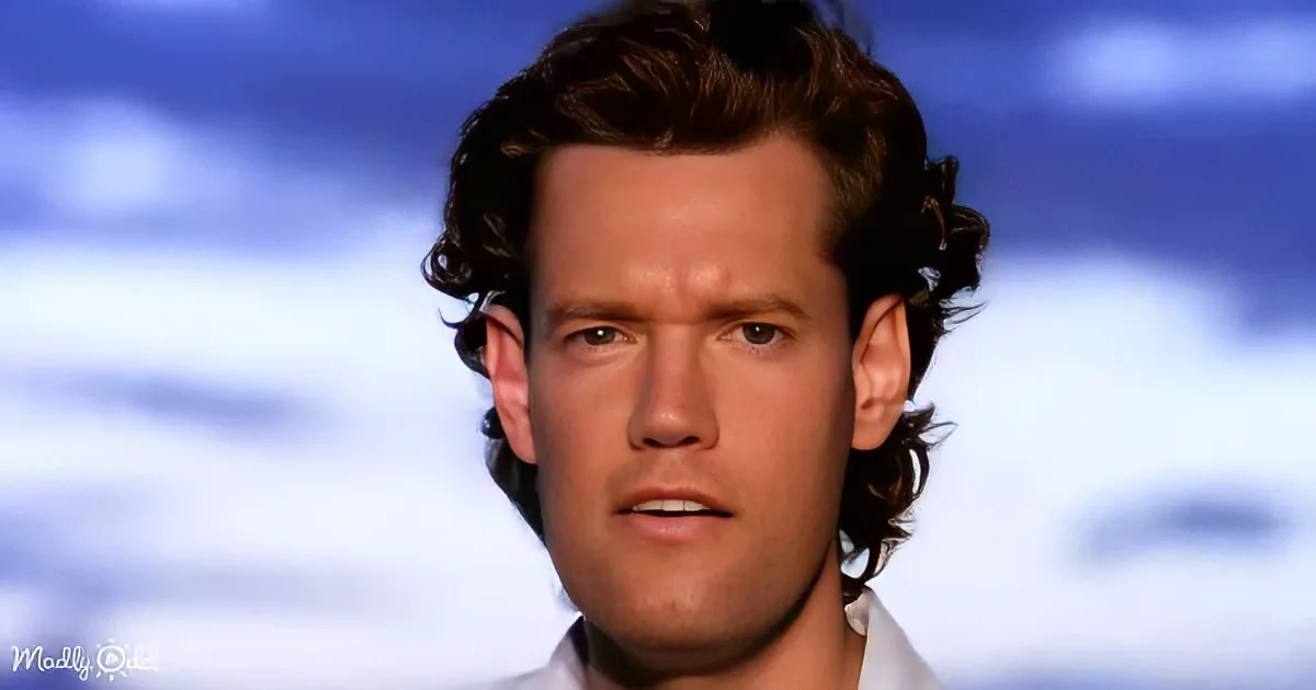 A man with curly hair and a white shirt stands against a cloudy sky backdrop.