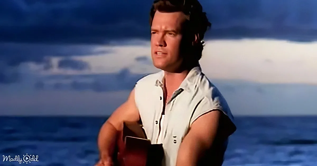A man in a white shirt holds a guitar, standing by the ocean under a cloudy sky.