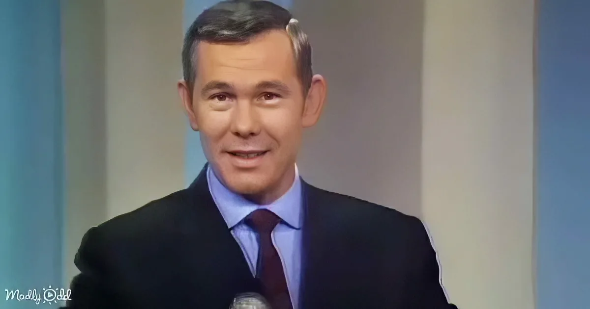 A man in a suit and tie smiles while holding a microphone, possibly on a TV show.