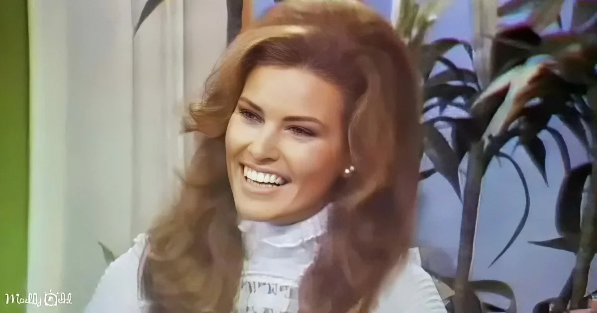 A smiling woman with long, wavy red hair wearing a white turtleneck, holding her hair back.