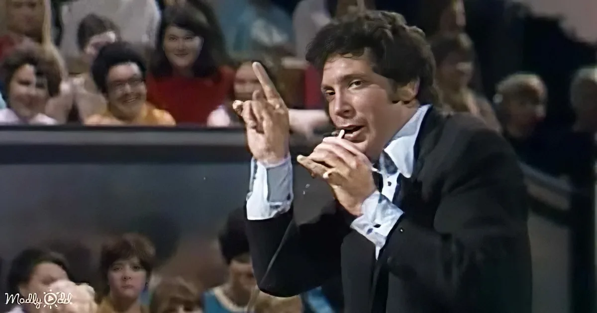 A man in a suit is speaking into a microphone, gesturing with his right hand.