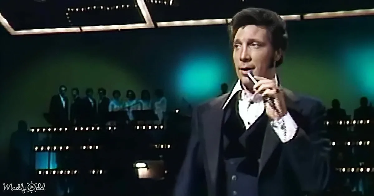 A man in a black suit and white shirt holds a microphone, singing on stage with a band in the background.