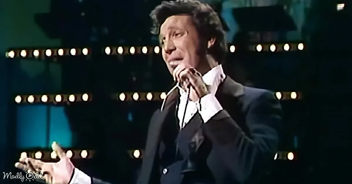A man in a suit sings passionately on stage with a microphone, surrounded by bright lights.