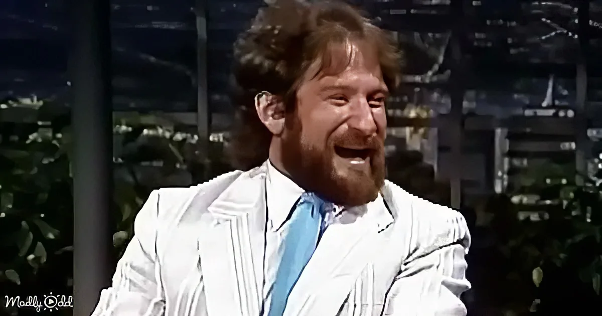 A man with a beard and white suit jacket is laughing, surrounded by greenery and a dark background.