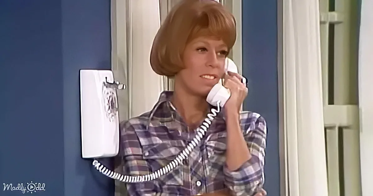 A woman in a plaid shirt talks on a white corded phone against a blue wall.