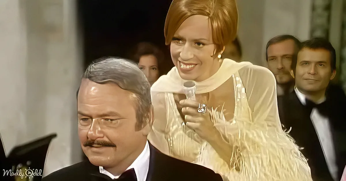 A woman in a white dress smiles while holding a microphone, standing behind a seated man in a tuxedo.