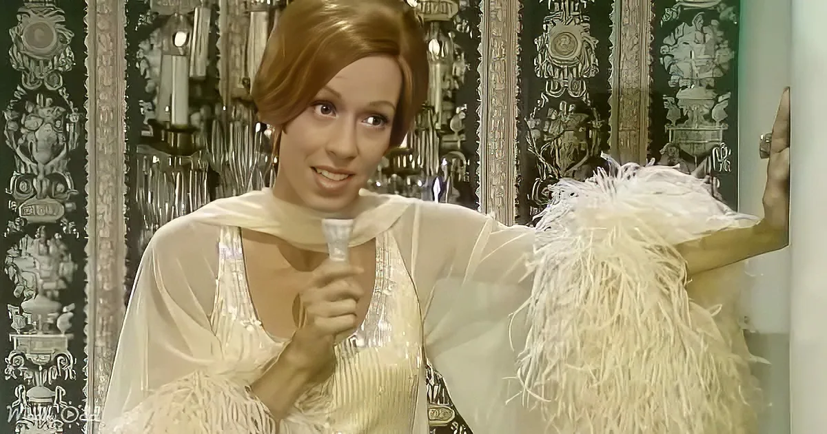 A woman in a white, fringed outfit holds a glass, smiling, with ornate, dark green and silver wall behind her.