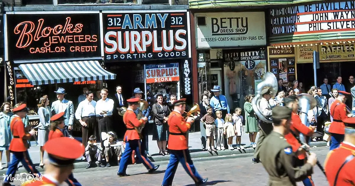 A marching band in red uniforms plays instruments on a street with spectators and vintage storefronts.