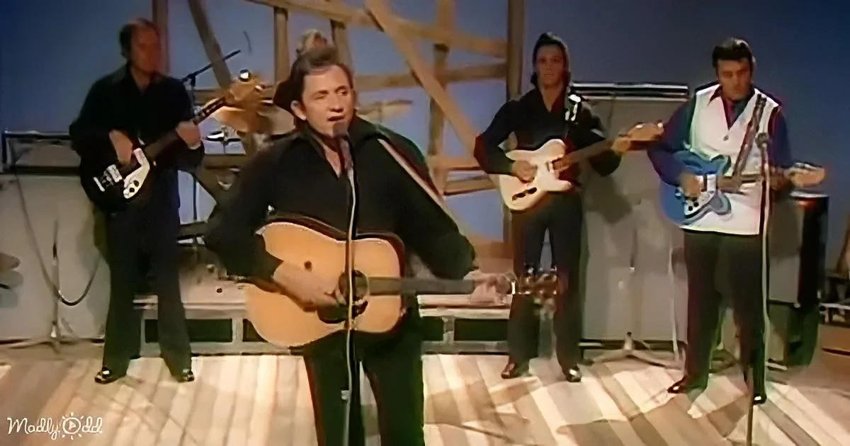 A man in a black jacket plays guitar and sings on stage with a band.