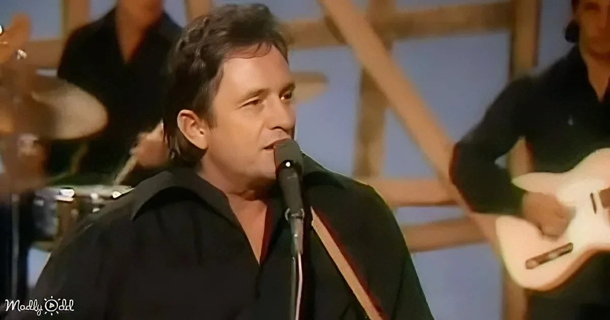 A man in a black shirt sings into a microphone, with a guitarist and drummer in the background.