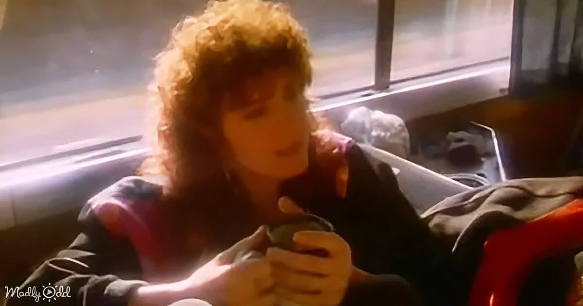 A person with curly hair sits in a car, holding a drink, with a window and sunlight visible.