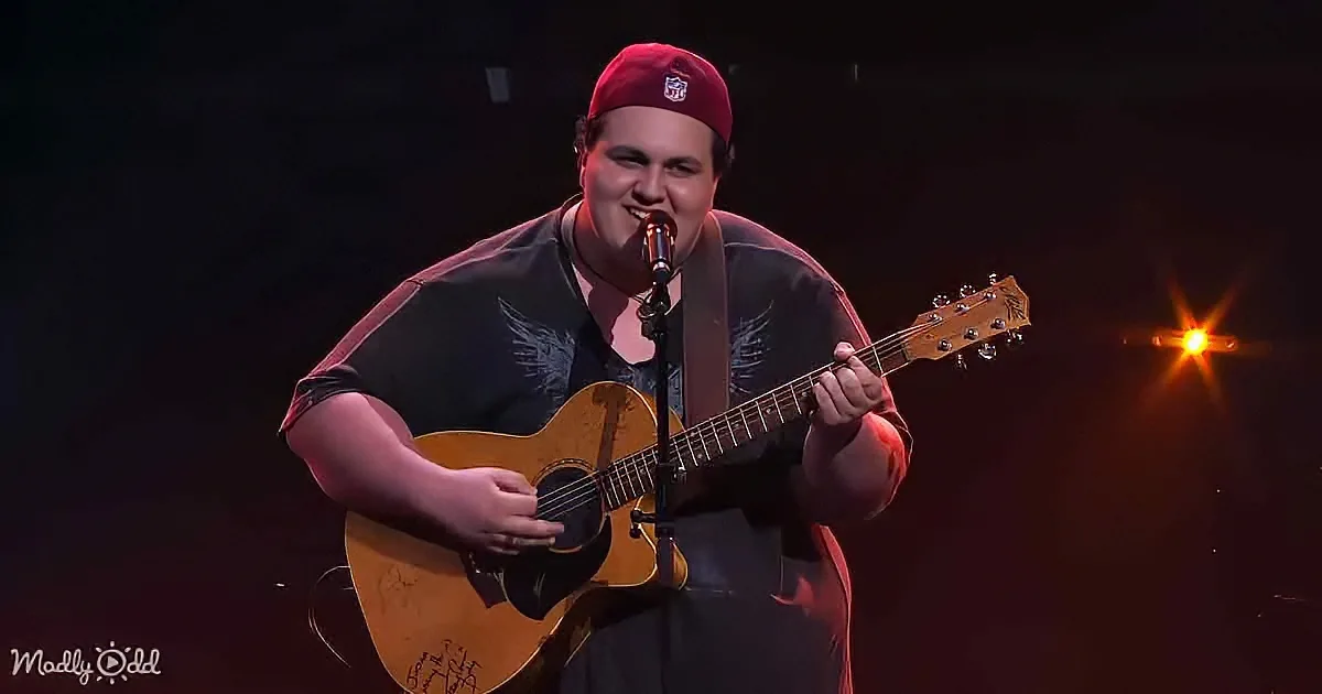 A man in a red cap plays an acoustic guitar on stage, singing into a microphone.