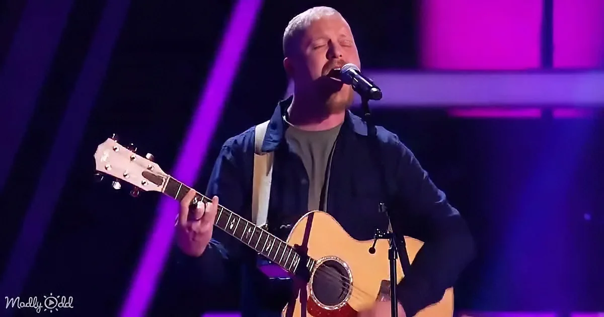 A man sings passionately while playing an acoustic guitar on stage under purple lighting.