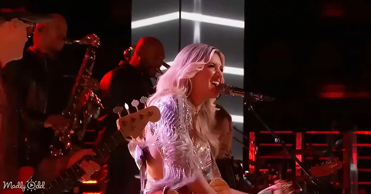 A woman with long, light-colored hair sings passionately into a microphone on stage, surrounded by musicians.