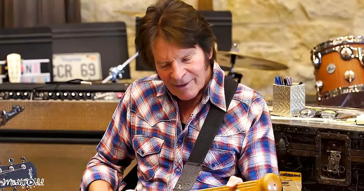 A man in a plaid shirt plays guitar in a music studio with amplifiers and drums.