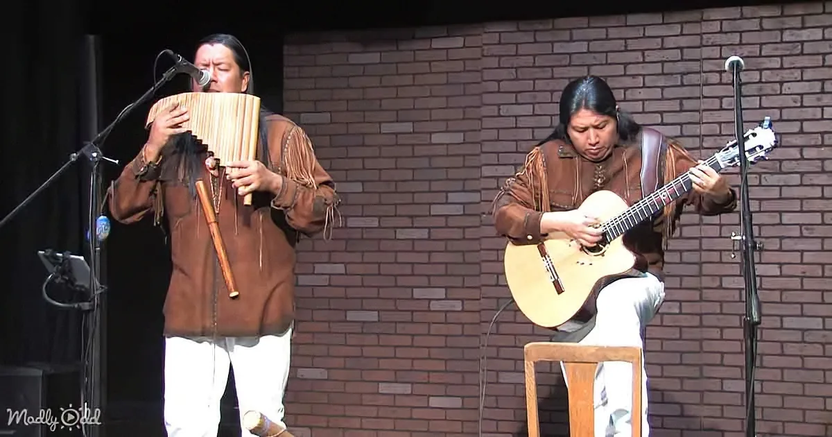A man plays a traditional instrument while another plays guitar on stage against a brick wall.