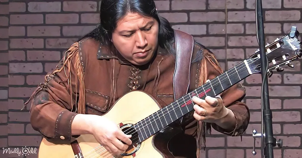 A person in a brown outfit plays an acoustic guitar against a brick wall backdrop.