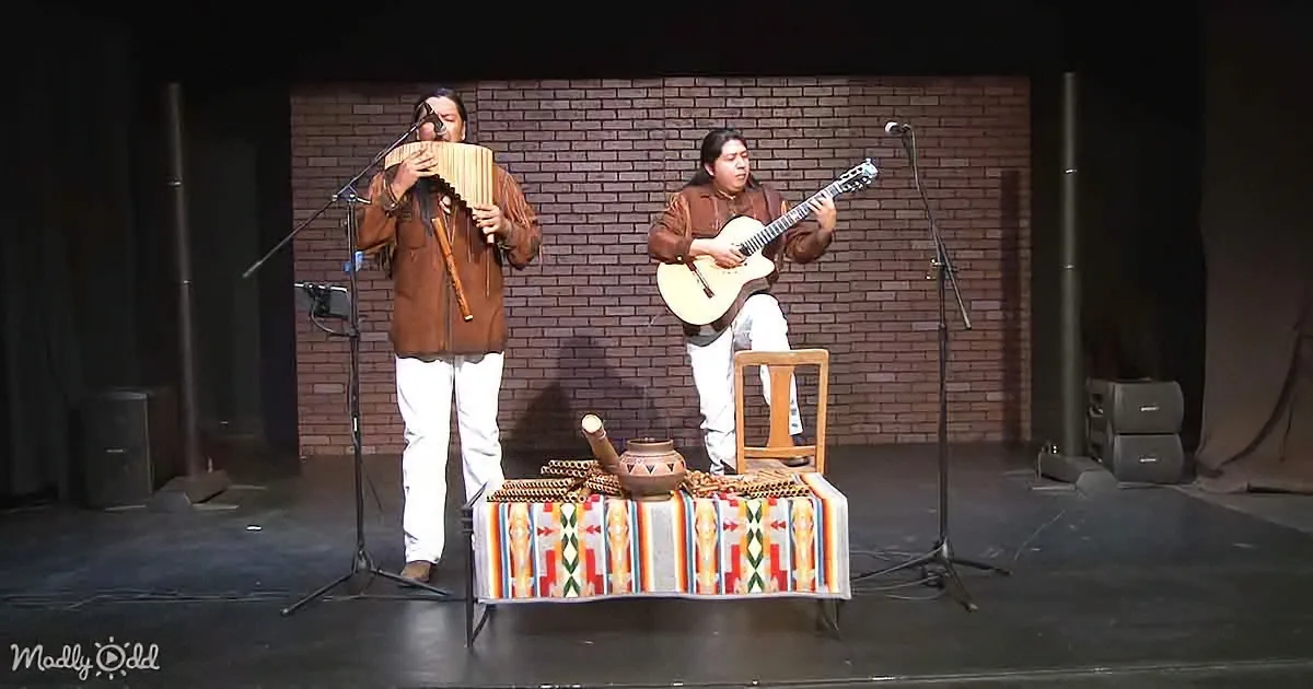 Two musicians perform on stage, one playing pan flute, the other guitar, with a colorful table in front.