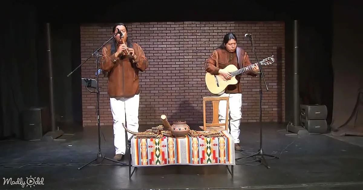 Two musicians perform on stage, one playing flute, the other guitar, with a table of Native American artifacts in front.